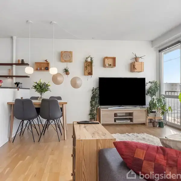 Stue med sofa og sofabord, hvor en stor fladskærm står på en hylde. Der er spisebord med fire stole ved siden af køkkenø. Planter og lamper dekorerer rummet.