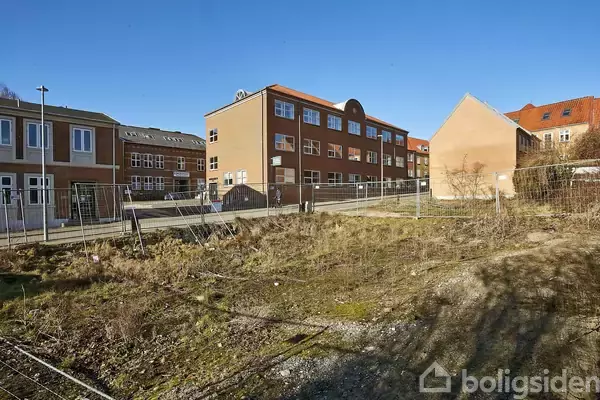 En åben grund omgivet af et hegn ligger i forgrunden, mens flere røde murstensbygninger ses i baggrunden under en klar blå himmel.