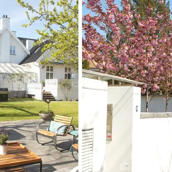Hvidt hus med skråt tag omkranset af grøn græsplæne og blomstrende træer. Terrassen har møbler og en lille plante. Lyseblåt himmel indikerer solrigt vejr.