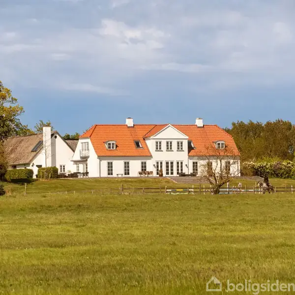 Ridebane med springopstillinger i brug, omgivet af grønne marker og en stor gårdbygning med rødt tag. På afstand ses flere bygninger og et åbent landskab under en klar himmel.