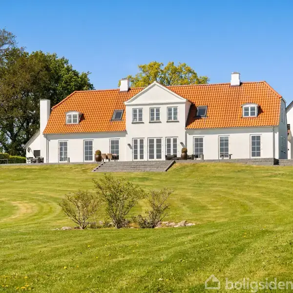 Et stort, hvidt hus med rødt tegltag står stille på en grøn mark omgivet af træer under en blå himmel.