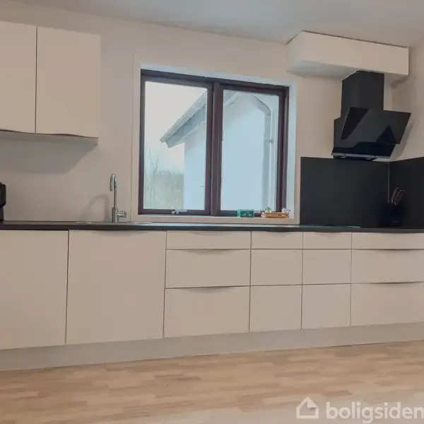 Et moderne køkken med hvide skabe står i et lyst rum med et stort vindue. Sort emhætte og bordplade tilføjer kontrast, mens trægulvet skaber varme.