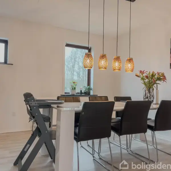 En spisestue har et hvidt bord omgivet af sorte stole. Over bordet hænger fem dekorative lamper. I baggrunden ses blomster, et billede på væggen og store vinduer.