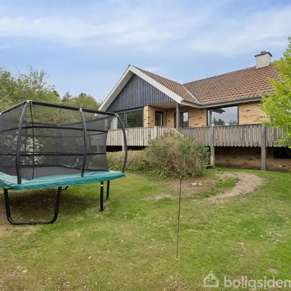 Trampolin i en grøn have foran et hus med træterrasse. Huset har skråt, rødt tag og omgives af buske og træer, der skaber en hyggelig atmosfære.