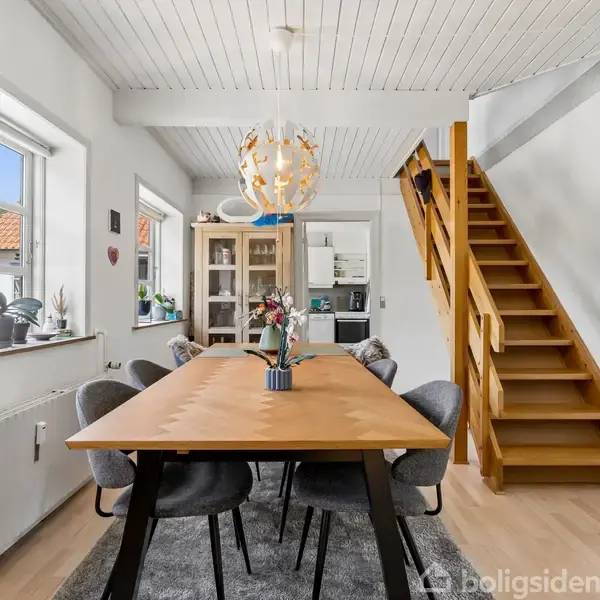 Træbord med seks polstrede stole. Over bordet hænger en moderne lampe. Rummet er lyst med trægulv, tæppe, trappe opadgående til højre, og et vindue til venstre.