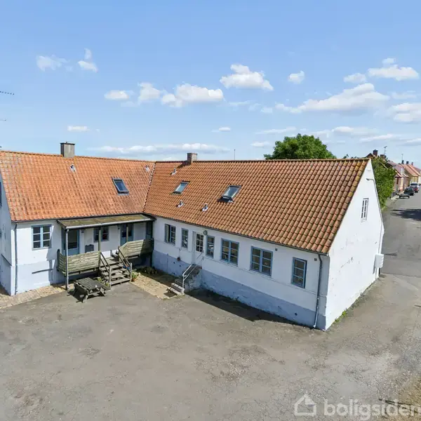 Hvid bygning med rødt tegltag står stille i en åben gård, omgivet af grusplads og grønne træer i et landligt område.