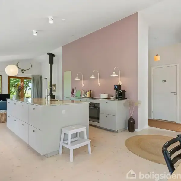 Et moderne køkken med en central ø og barstol, omgivet af stue med blå sofa. Vinduer viser grøn have udenfor, og væggen er malet lilla.