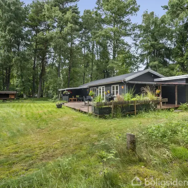 Sort træhus står på en græsplæne omgivet af skov. På verandaen ses blomsterpotter og havemøbler, mens et skur er placeret i baggrunden blandt træerne.