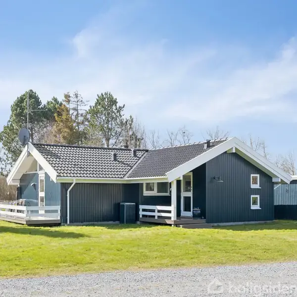 En mørk træhytte med lys tag tager fokus, omgivet af en grøn græsplæne og træer. Hytten har en terrasse og ligger i et åbent, naturskønt område.