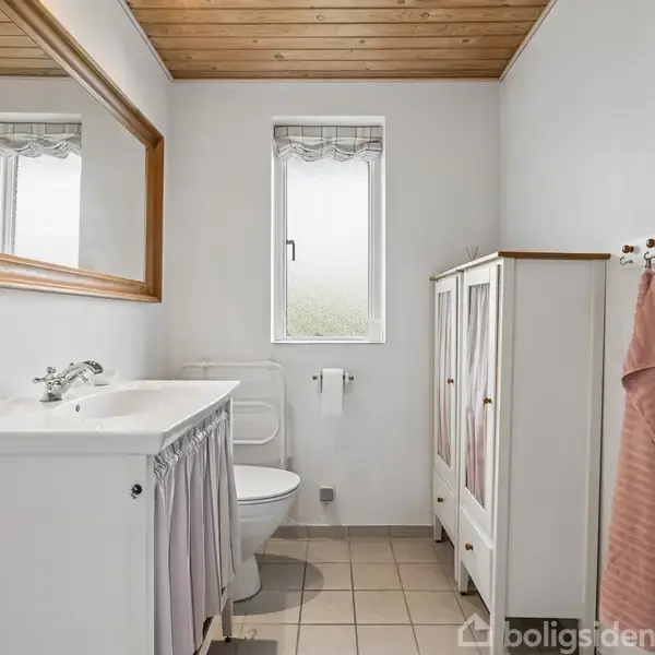 Badeværelse med hvid vask på et hvidt skab, et spejl over vasken, og et vindue i baggrunden. Der er også et hvidt skab og en lyserød håndklæde på væggen.