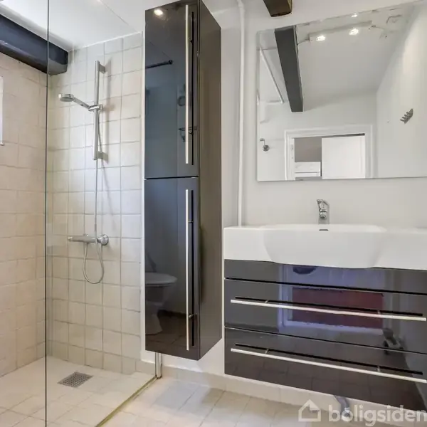 Håndvask med skuffemøbel og spejl over. Brusekabine til venstre med glasvæg og hvidflisevæg. Moderne, lyst badeværelse med vindue, hvid gulvflise og smalle, mørke skabe.