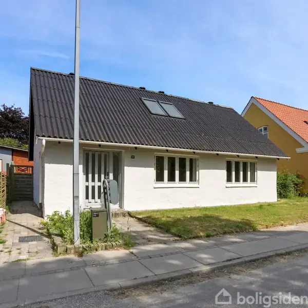En hvid, etplans villa står stille i et åbent boligområde. Huset har sort tag og store vinduer. Ved siden af huset er en bygningsgul villa. Der er græsplæner og fortov foran.