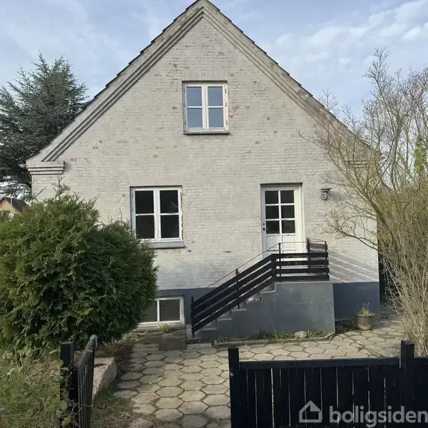 Et hvidt murstenshus med en sort trappe ind mod indgangen. Omkranset af en have med buske og flisebelagt sti fører op til huset.