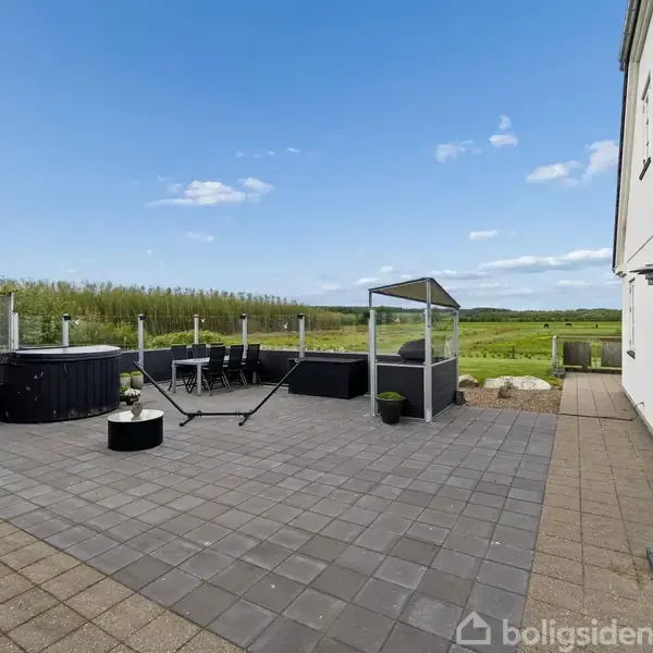 Hot tub står på en flisebelagt terrasse ved siden af havebord og stole, omgivet af glashegn. I baggrunden er der udsigt over grønne marker og blå himmel.