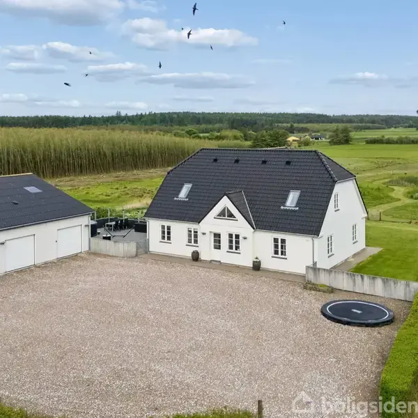 Et hvidt hus med sort tag står ved siden af en garage, omgivet af marker. En trampolin er nedgravet ved grusgården, og fugle flyver over det åbne landskab.
