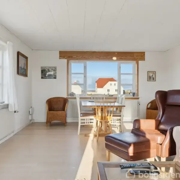 Stue med lænestol og sofabord, der har en fjernbetjening og briller. I baggrunden findes et spisebord med stole nær vinduer med udsigt til et hus udenfor.