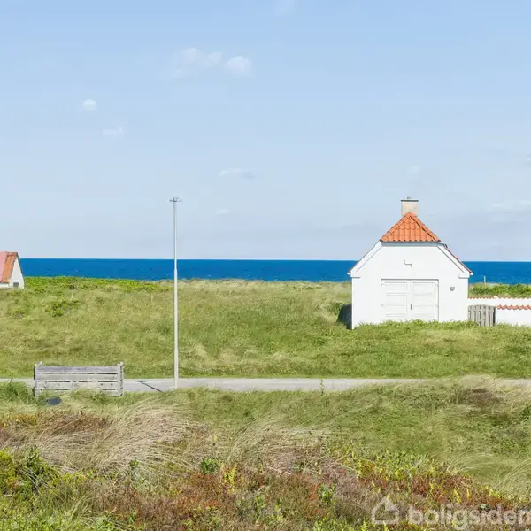 To hvide huse med røde tage står stille i en græsbeklædt klitlandskab, med en træbænk i forgrunden, mens en blå horisont af hav udfolder sig i baggrunden.