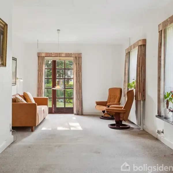 En stue med en brun sofa og en lænestol står mod vinduer og glasdør, der fører ud til en have. Gardiner hænger ved siden af vinduerne.