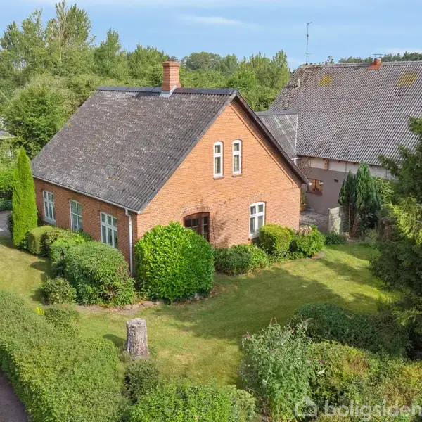 Et rødt murstenshus med skråt sort tag står omgivet af en velholdt grøn have med buske og træer. En asfalteret vej findes til venstre for huset i et grønt nabolag.