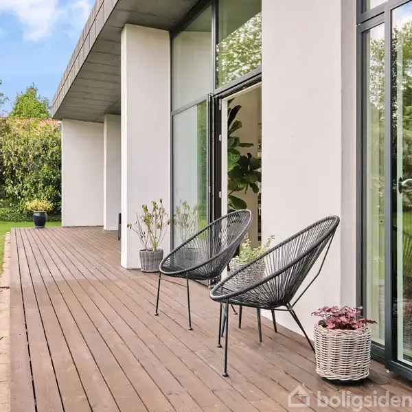 To moderne stole står på en træterrasse foran store glasvinduer i et hus, omgivet af en frodig have med græs og buske.