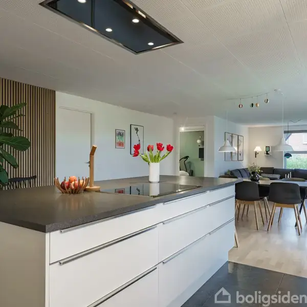 Køkkenø med hvid skabsløsning, dekoreret med en vase med røde tulipaner. Åben stue i baggrunden med spisebord, stole, og grønne planter, giver et moderne og lyst indtryk.