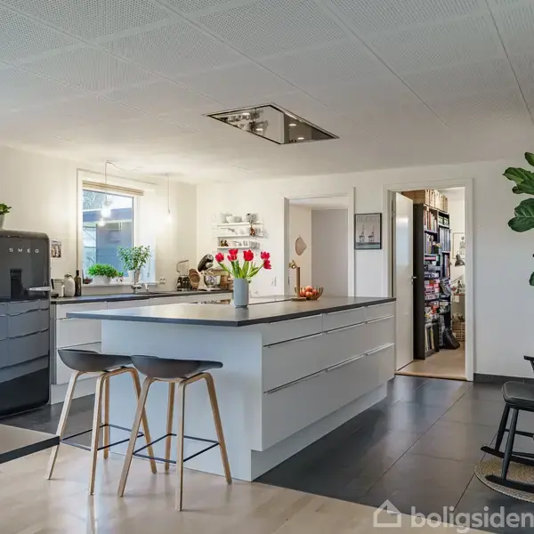 Et moderne køkken med en stor køkkenø, to barstole, og høje skabe. Køleskab, planter og blomster tilføjer farve, mens en gang leder til et lagerområde.