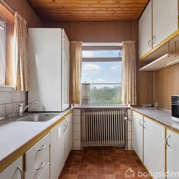 Et køkken med hvide skabe og en brun bordplade. En mikrobølgeovn står på bordet. Vinduet i baggrunden har udsigt til træer. Radiator under vinduet. Ingen tekst.