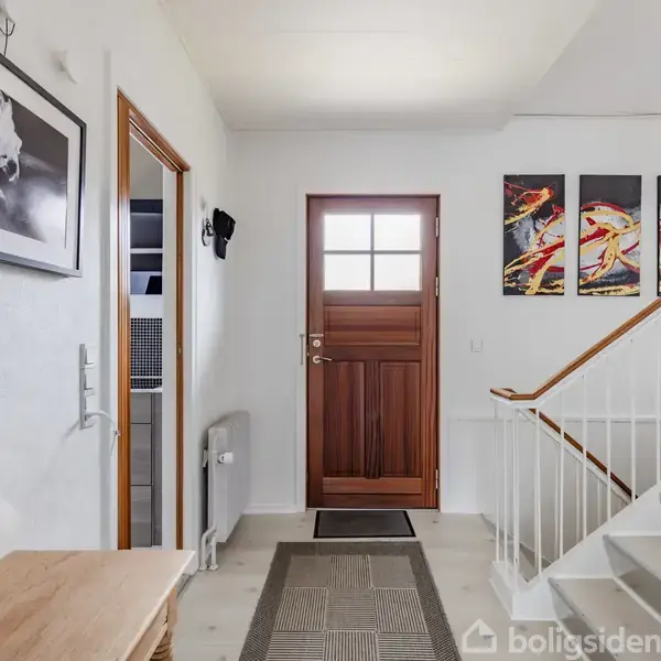 Dør i træ fører ind til rummelig entré med lyse vægge. Moderne kunst hænger over hvid trappe. Sort-hvidt fotografi er placeret over et lille bord ved siden af døren.