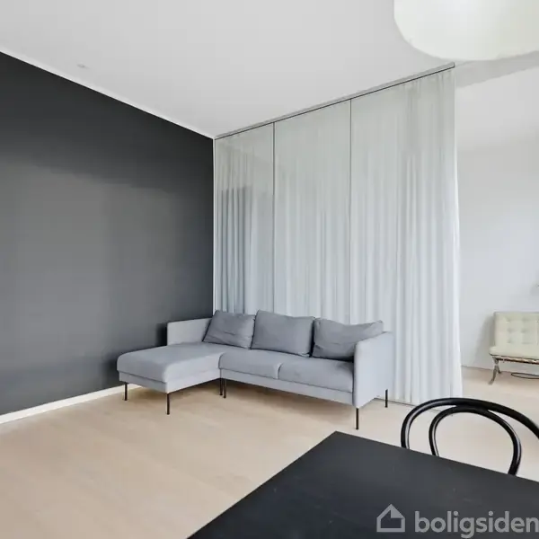 Grå sofa står mod en sort væg i en lys stue. Rummet har en beige stol, et bord med stol og store vinduer med gardiner.