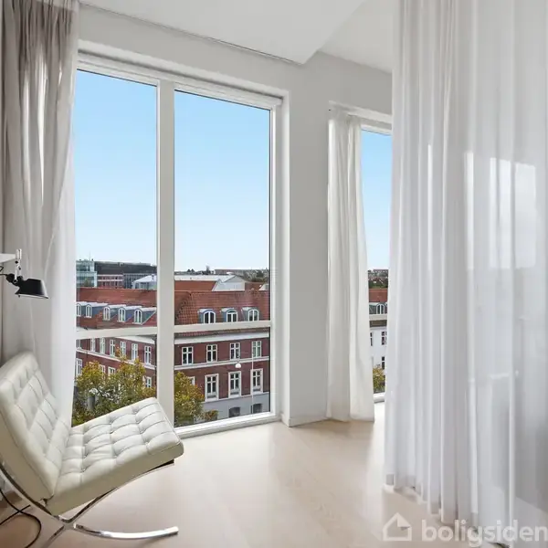 En hvid lænestol står ved siden af et stort vinduesparti, hvor lyse gardiner hænger. Udsigten viser røde hustage og træer under en klar blå himmel.