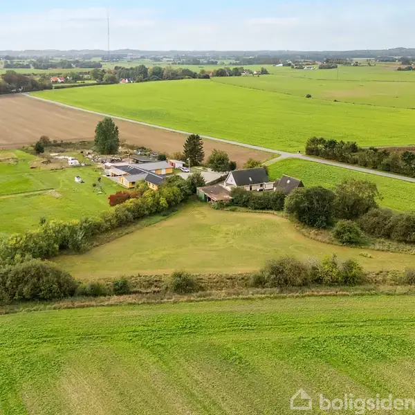 Luftfoto af en gård med flere bygninger omgivet af marker og træer. Veje snor sig gennem det landlige landskab med marker, der strækker sig mod horisonten under en blå himmel.