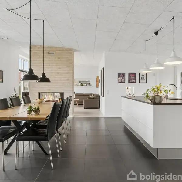 Spisebord med sorte stole står i et moderne køkkenalrum med hvidt interiør, hængelamper og et langbord. I baggrunden er en stue med sofa og pejs.