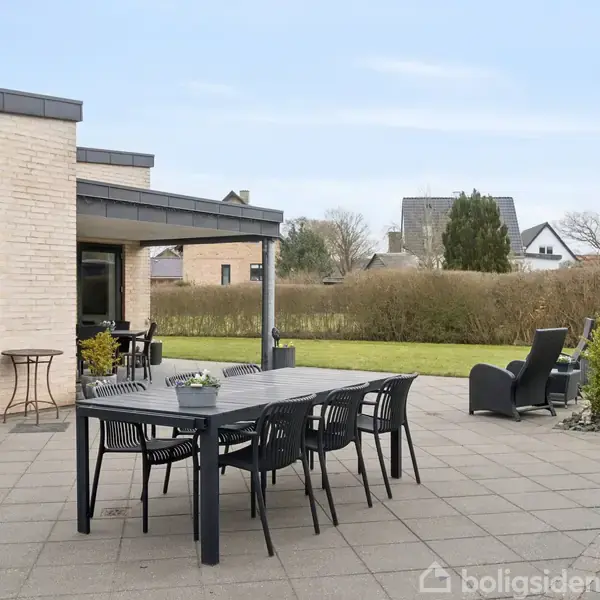 Udendørs terrasse med sort spisebord og seks stole placeret på fliser, omkranset af et moderne husmur og have. Enkelte havemøbler og små buske i baggrunden.