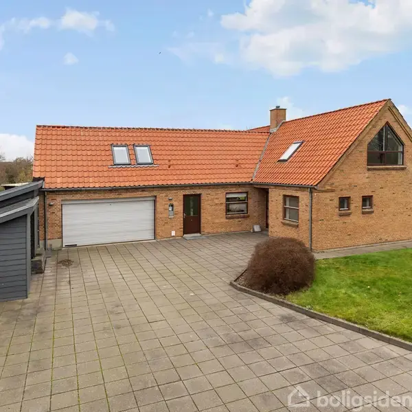 Et murstenshus med rødt tegltag står ved en flisebelagt indkørsel; en græsplæne omkranser huset, der er placeret i et villakvarter med træer i baggrunden.