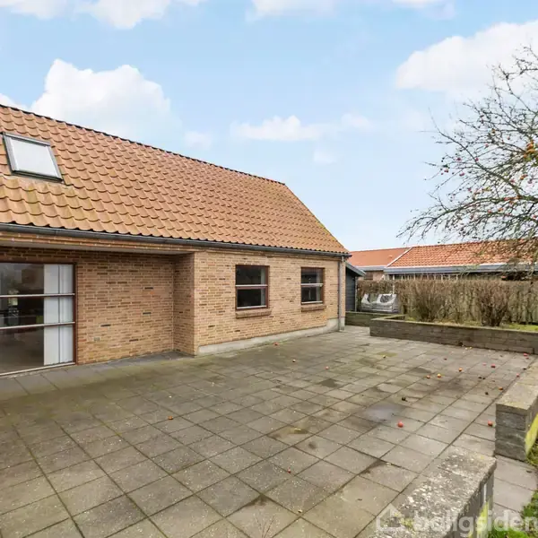 Et murstenshus med rødt tegltag og store vinduer står ved en flisebelagt terrasse. Til højre ses et bar træ og en lav mur, omgivet af en have.