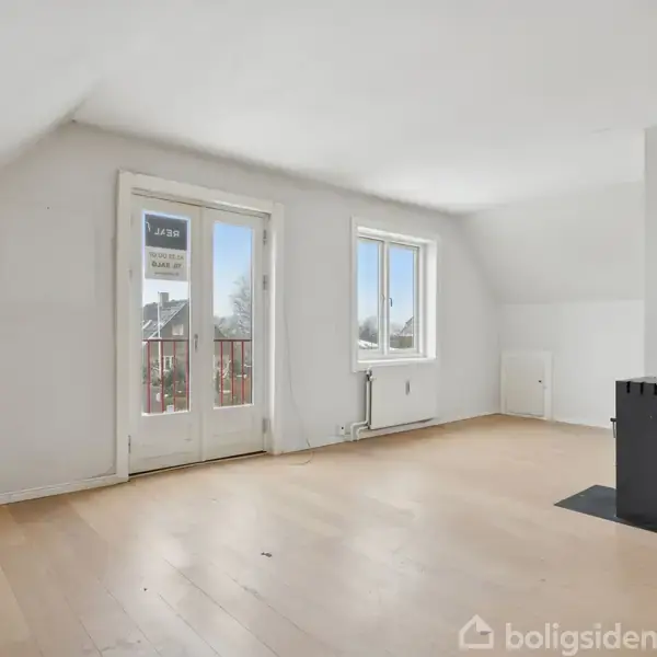 Et tomt stueområde med et fritstående sort brændeovn til højre. Der er lyse træplanker på gulvet og et vindue samt glasdør til en balkon. Skiltet "Til salg" ses.