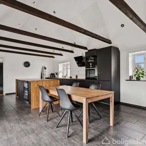 Et køkken med træbjælker i loftet, træbord og sorte stole. Et sort køkkenmodul står til højre. Der er lyse gulve, vinduer og en dør i baggrunden.