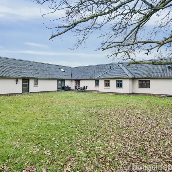 Hus med fladt tag står stille i en åben have omgivet af grønne græsarealer og nøgne trægrene. Skydedør fører til terrassen med sorte havestole.