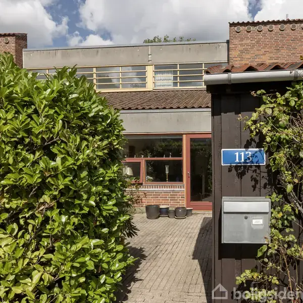 En postkasse med nummeret 113 er monteret på en sort væg ved siden af en grøn busk. Bygningen i baggrunden har store vinduer og en flisebelagt indgang.