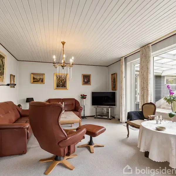En stue med brune lædermøbler og et rundt bord med dug, placeret ved et stort vindue. Væggene er dekoreret med malerier, og der er en lysekrone i loftet.