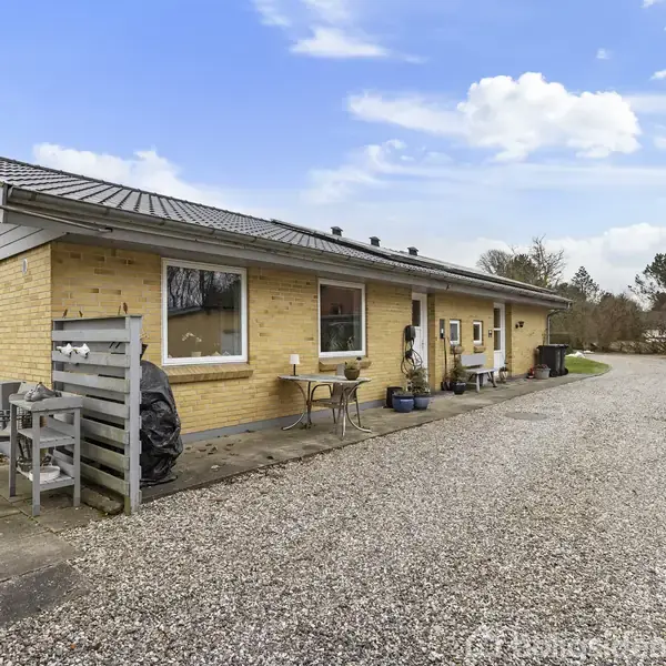 Gulstenshus står stille; grusvej foran; træer i baggrunden. Bord og stole i gårdhave til venstre. Klar blå himmel med skyer.