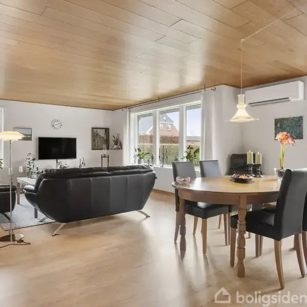 En stue med sorte lædersofaer står foran et tv på en stander; et spisebord med fire stole til højre. Rummet har lyse vægge, trægulve og store vinduer.