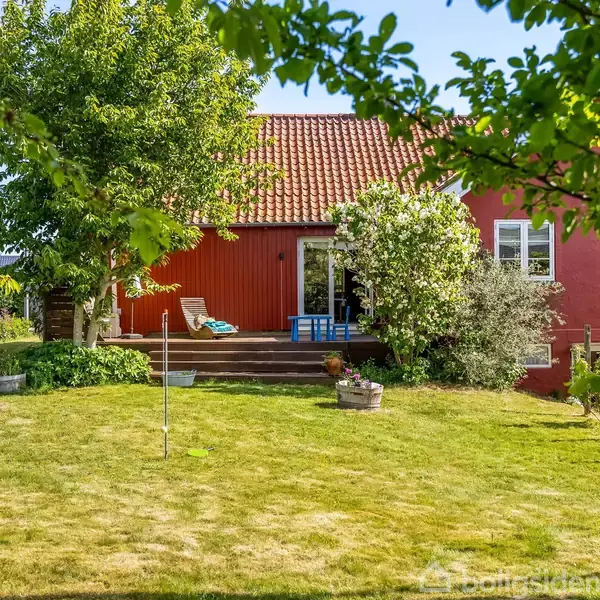 Et rødt hus med tagterrasse, omgivet af en frodig have med træer og græsplæne. En liggestol står ved terrassen, og omgivelserne fremstår solrige og rolige.