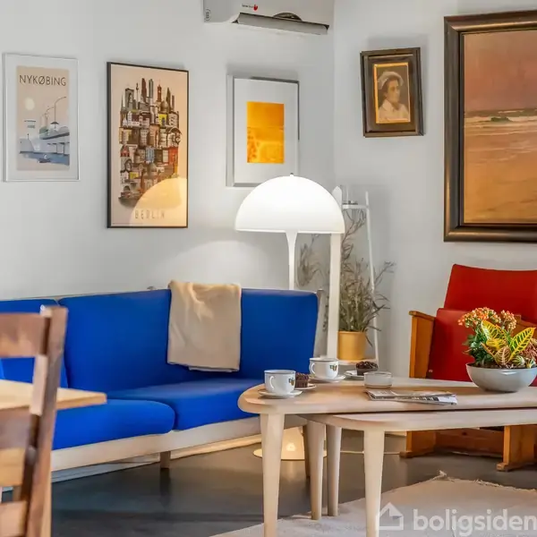 Blå sofa med tæppe står i en stue, omgivet af kunstplakater og malerier på væggene. Rundt sofabord med bøger, kopper og en skål med planter. Skilte med "NYKØBING".