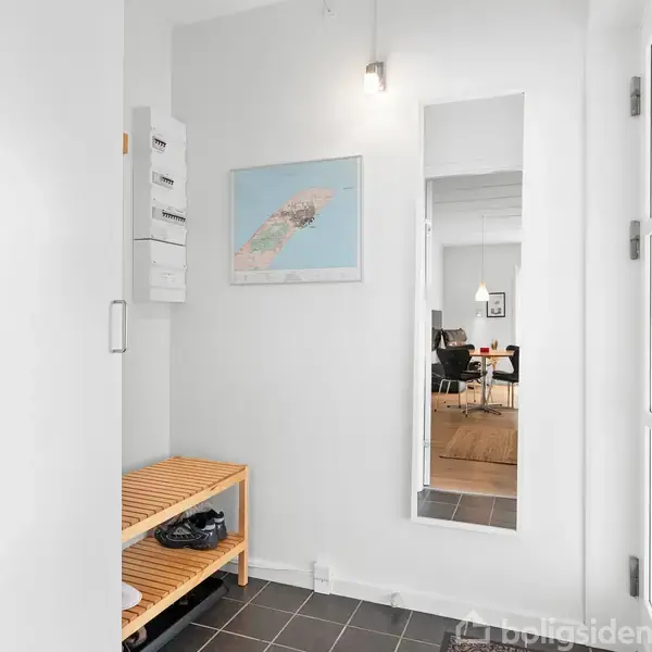 En lys lille entré med en træbænk til sko i venstre side, en vægmonteret spejl og kort samt fliser på gulvet. Det leder til en spisestue gennem en åbning.