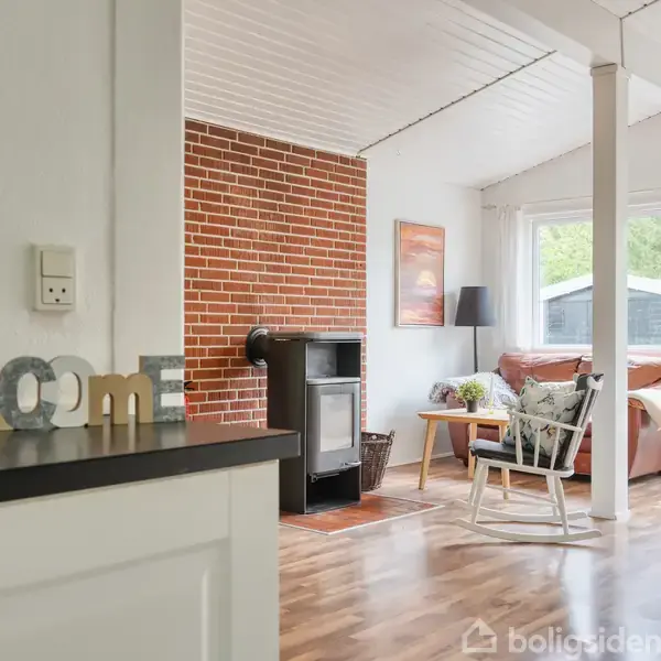 Et sortmalet træhus står stille, omringet af græs og træer. Der er en terrasse med havemøbler, der inviterer til udendørs ophold.