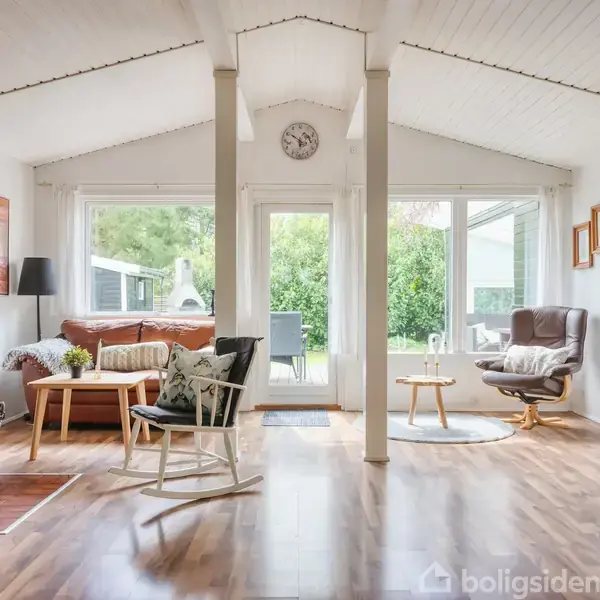 Stue med brændeovn ved murstensvæg, stående ved siden af et bord og stol. Hyggelig atmosfære med lysindfald gennem vinduer. Tekst på reolen: "wElcOmE". Hvide vægge og loft.