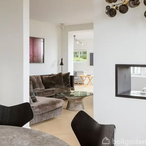 En blød, brun sofa er placeret i en moderne stue ved siden af et rundt glasbord. I baggrunden ses et stort vindue og lysindfald i et åbent rum.