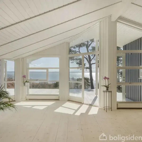 Et lyst rum med store vinduer, der giver udsigt til en skov og havet. En plante og en blomsterpotte står ved vinduet. Højt, hvidt loft skaber en rummelig atmosfære.