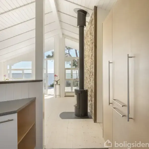 Et sort træhus med rødt tag står ved vandet. Huset har store vinduer, og der er en grusbelagt indkørsel og græsplæne omkring det, med et hav i baggrunden.